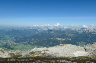 A panoramic view from an Alpine top on a vast valley. There are sharp mountains and high peaks around. The Alpine slopes are almost barren. Lush green valley. Bright day. Serenity and freedom.