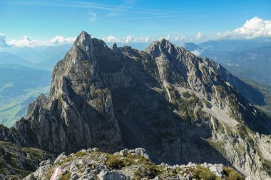 A panoramic view from an Alpine top on a vast valley. There are sharp mountains and high peaks around. The Alpine slopes are almost barren. Lush green valley. Bright day. Serenity and freedom.