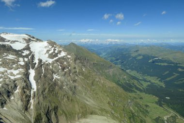 Panoramic view from top of Hohe Sonnblick in Austrian Alps on Grossglockner. The whole area is partially covered with snow, lush green meadows below. Many mountain chains in the back. Sunny day.