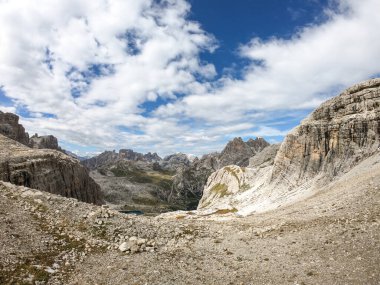 A panoramic view on Dolomites in Italy. There are sharp and steep mountain slopes around. Lots  of lose stones and pebbles. The sky is full of soft clouds. Raw landscape. Serenity and calmness