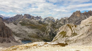 A panoramic view on Dolomites in Italy. There are sharp and steep mountain slopes around. At the bottom of a small valley there is a small navy blue lake. The sky is full of soft clouds. Raw landscape