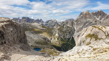 A panoramic view on Dolomites in Italy. There are sharp and steep mountain slopes around. At the bottom of a small valley there is a small navy blue lake. The sky is full of soft clouds. Raw landscape