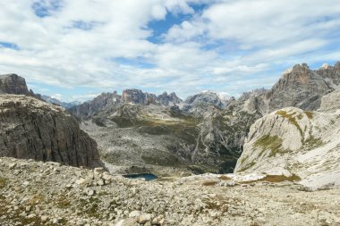 A panoramic view on Dolomites in Italy. There are sharp and steep mountain slopes around. At the bottom of a small valley there is a small navy blue lake. The sky is full of soft clouds. Raw landscape