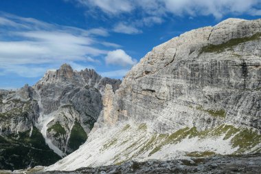 A panoramic view on Dolomites in Italy. There are sharp and steep mountain slopes around. Lots  of lose stones and pebbles. The sky is full of soft clouds. Raw landscape. Serenity and calmness