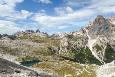 A panoramic view on Dolomites in Italy. There are sharp and steep mountain slopes around. At the bottom of a small valley there is a small navy blue lake. The sky is full of soft clouds. Raw landscape