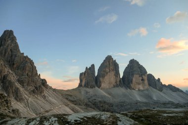 A panoramic capture of the sunset above Tre Cime di Lavaredo (Drei Zinnen) and surrounding mountains in Italian Dolomites. The mountains are surrounded by pink and orange clouds. Golden hour. Serenity