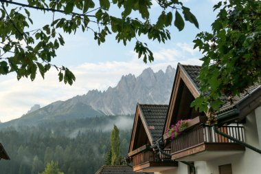 A row of houses with the view on high and sharp Dolomites in Italy. There is a dense forest between the houses and the mountains, partially shrouded in morning haze. Few tree branches above