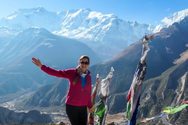 A woman in hiking outfit standing on a barren pathway along Annapurna Circuit Trek. There are few prayer flags next to her. Snow caped Annapuna chain in the back. Achievement and completion