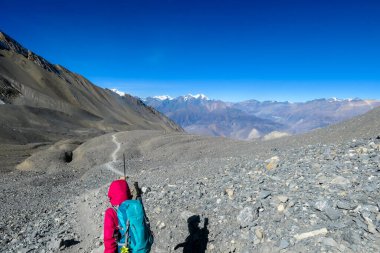 A woman hiking through dry path in Himalayan valley, located in Mustang region, Annapurna Circuit Trek in Nepal. She is going down a steep slope. She carries a heavy backpack. Harsh landscape.