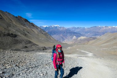 A woman hiking through dry path in Himalayan valley, located in Mustang region, Annapurna Circuit Trek in Nepal. She is going down a steep slope. She is smiling friendly. Harsh landscape.