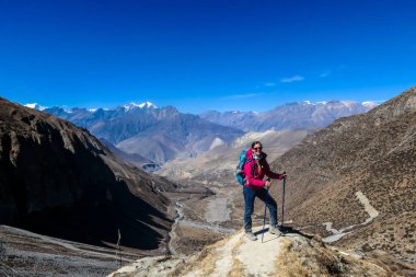 A woman hiking through dry path in Himalayan valley, located in Mustang region, Annapurna Circuit Trek in Nepal. She is having a short break, supporting herself on hiking sticks. Harsh landscape.