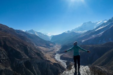 A woman wearing a beanie and blue jumper, spreads her arms wide, breathing deeply the fresh mountain air. Freedom and happiness. Below Manang valley stretches in Himalayas, along Annapurna Circuit.