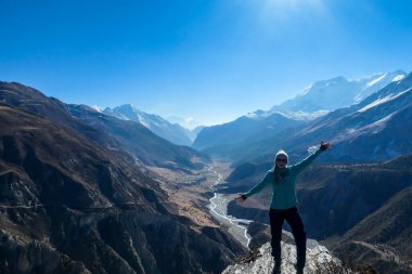 A woman wearing a beanie and blue jumper, spreads her arms wide, breathing deeply the fresh mountain air. Freedom and happiness. Below Manang valley stretches in Himalayas, along Annapurna Circuit.