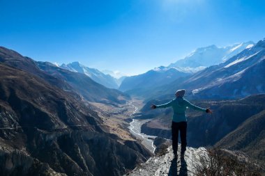 A woman wearing a beanie and blue jumper, spreads her arms wide, breathing deeply the fresh mountain air. Freedom and happiness. Below Manang valley stretches in Himalayas, along Annapurna Circuit.