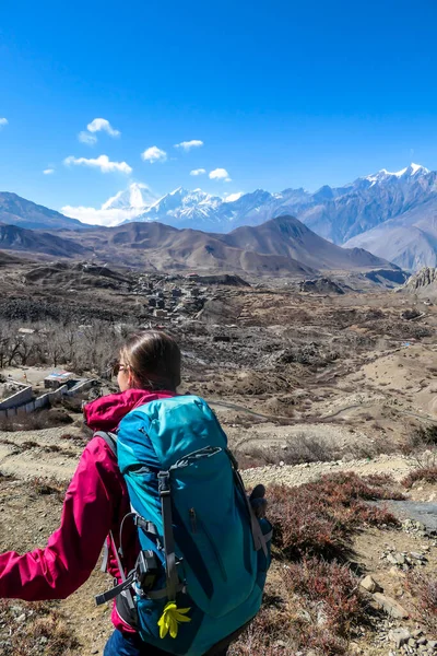 A woman hiking through Himalayan valley, located in Mustang region, Annapurna Circuit Trek in Nepal. She is having a short break, supporting on hiking sticks and enjoying the view on small village