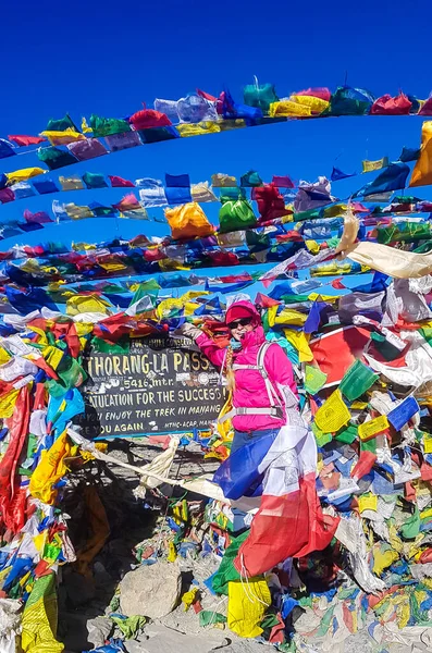 A woman standing between prayer flags at the top of Thorung La Pass, Annapurna Circuit Trek, Nepal. She is very happy. Colorful prayer flags attached to the stone wall, blown by the wind. Clear sky.