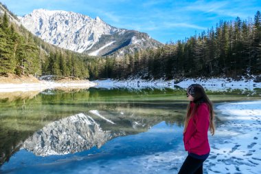 A woman walking around the shore of Green Lake, Austria. Powder snow covering the mountains and ground. Soft reflections of Alps in calm lake's water. Winter landscape of Austrian Alps. Calmness