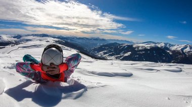 A woman lying in powder snow any enjoying the sun on top of Katschberg in Austria. She is happy and joyful. Panoramic view on the surrounding mountains. Winter wonderland, Clear and sunny winter day