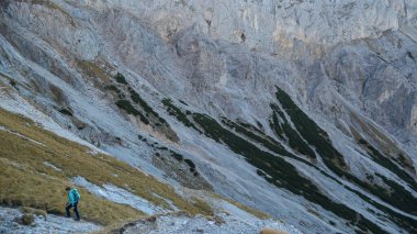 A woman hiking between sharp and steep slopes of Hochschwab in Austrian Alps. The massive stony wall rockets up very steep. The flora overgrowing the slopes is turning golden. Autumn vibes. Wilderness