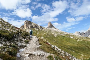 A woman with backpack and sticks hiking on a narrow path in Italian Dolomites. There are sharp and steep mountains. At the bottom of a small valley there is a small, navy blue lake. Raw landscape.