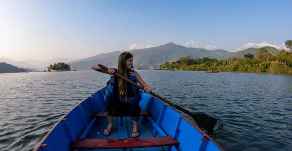 A woman in blue safety jacket, sitting in a blue boat and paddling across Phewa Lake in Pokhara, Nepal. Behind her there are high, snow capped Himalayas with Mt Fishtail (Machhapuchhare) between them