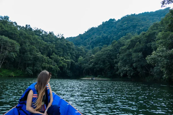 A woman sitting in a blue boat and enjoying a tour across Phewa Lake in Pokhara, Nepal. Behind her there is a dense forest. The woman is enjoying a peaceful ride. Relaxation and chill