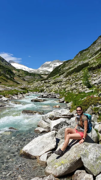 Woman wearing a backpack enjoying a cascading waterfall in the Alps in Ankogel group in Austria. The rushing torrent is coming from a glacier water. High mountains around, overgrown with small bushes