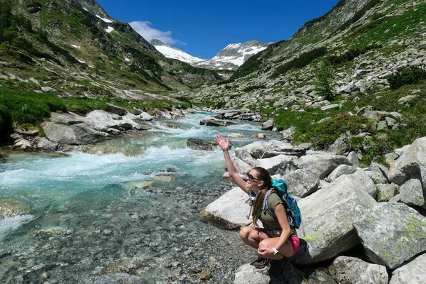 Woman wearing a backpack enjoying a cascading waterfall in the Alps in Ankogel group in Austria. The rushing torrent is coming from a glacier water. High mountains around, overgrown with small bushes