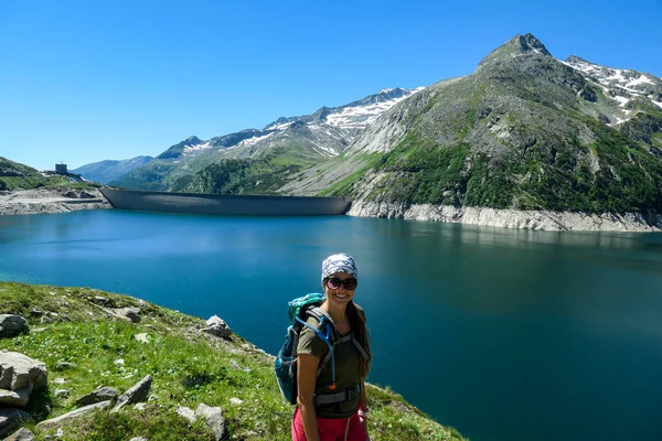 A woman with hiking backpack standing at the side of a lake at Koelnbreinsperre dam in Austria. The lake has navy blue color. High Alps around. There is a glacier in the back. Adventure and discovering