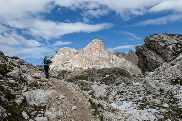 A woman with backpack and sticks hiking on a narrow path in Italian Dolomites. There are sharp and steep mountains. At the bottom of a small valley there is a small, navy blue lake. Raw landscape.