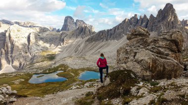 Woman hiking with the view on small, navy blue lakes at the bottom of the valley in Italian Alps. The lakes are surrounded by high and steep peaks The sky is full of soft clouds. Raw landscape. Remedy