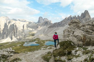 Woman hiking with the view on small, navy blue lakes at the bottom of the valley in Italian Alps. The lakes are surrounded by high and steep peaks The sky is full of soft clouds. Raw landscape. Remedy