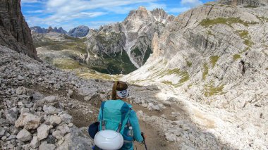 A woman with backpack and sticks hiking on a narrow path in Italian Dolomites. There are sharp and steep mountains around her. Lots of lose stones. Raw and desolated landscape. Following the pathway