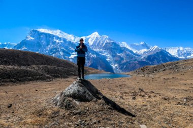 A young man wearing a blue jacket, standing on a huge rock next to Ice Lake, Annapurna Circuit Trek, Himalayas, Nepal. High, snow caped Annapurna chain in the back. Solitude and awareness.