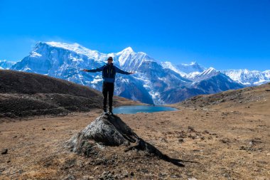 A young man wearing a blue jacket, standing on a huge rock next to Ice Lake, Annapurna Circuit Trek, Himalayas, Nepal. High, snow caped Annapurna chain in the back. Solitude and awareness.
