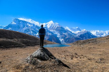 A young man wearing a blue jacket, standing on a huge rock next to Ice Lake, Annapurna Circuit Trek, Himalayas, Nepal. High, snow caped Annapurna chain in the back. Solitude and awareness.