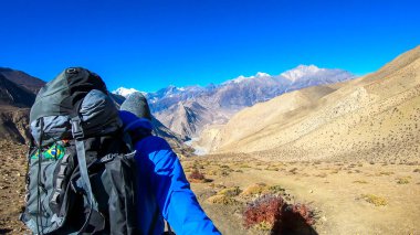 A man hiking through dry paths in Himalayan valley, located in Mustang region, Annapurna Circuit Trek in Nepal and taking a selfie. The man carried a heavy backpack. Barren slopes. Harsh landscape.