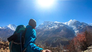 A man hiking and taking a selfie with snow caped Annapurna chain in the back, Annapurna Circuit Trek, Himalayas, Nepal. High mountains around. The man is admiring the landscape. Serenity and calmness.