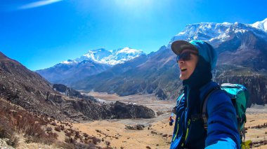 A man hiking and taking a selfie with snow caped Annapurna chain in the back, Annapurna Circuit Trek, Himalayas, Nepal. High mountains around. The man is a bit tired. Serenity and calmness.