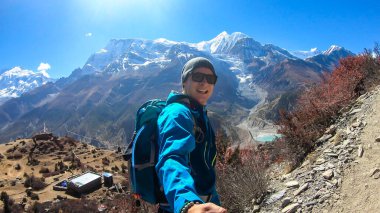 A man hiking and taking a selfie with snow caped Annapurna chain in the back, Annapurna Circuit Trek, Himalayas, Nepal. High mountains around. The man is admiring the landscape. Serenity and calmness.