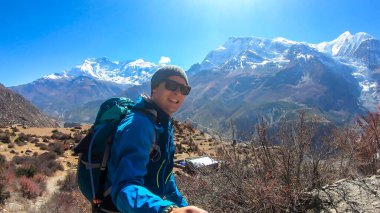 A man hiking and taking a selfie with snow caped Annapurna chain in the back, Annapurna Circuit Trek, Himalayas, Nepal. High mountains around. The man is admiring the landscape. Serenity and calmness.