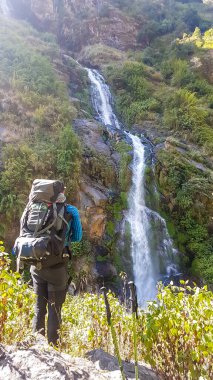 A man with a big hiking backpack standing in front of a tall waterfall in Himalayas, along Annapurna Circuit Trek, Nepal. The waterfall falls on the sharp, rocky slopes. Power of the nature.