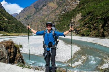 A man wandering along the slopes of Annapura Circuit Trek in Nepal. There is a strong torrent behind him. He is carrying a big backpack. A few high mountain chains in the back. Himalayan valley.