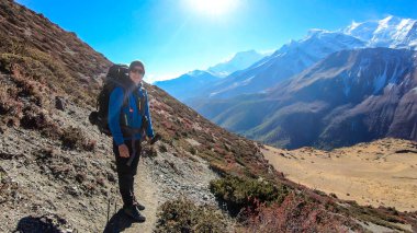 Man with big hiking backpack trekking along Annapurna Circus in Himalayas, Nepal, with the view on Annapurna Chain. Dry and desolated landscape. High, snow capped mountain peaks. Happiness and freedom