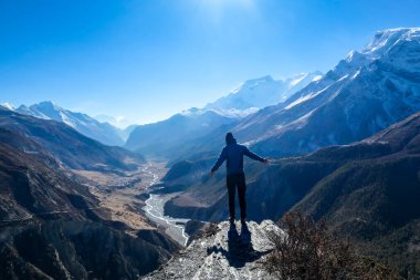 A man wearing a beanie and blue jumper, stands at the rock edge, breathing deeply the fresh mountain air. Freedom and happiness. Below Manang valley stretches in Himalayas, along Annapurna Circuit.