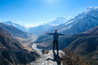 A man wearing a beanie and blue jumper, spreading his arms wide, breathing deeply the fresh mountain air. Freedom and happiness. Below Manang valley stretches in Himalayas, along Annapurna Circuit.