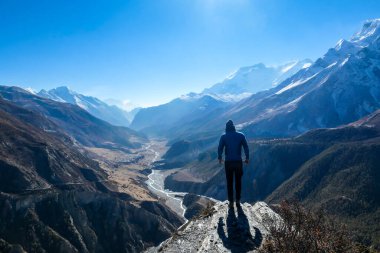 A man wearing a beanie and blue jumper, stands at the rock edge, breathing deeply the fresh mountain air. Freedom and happiness. Below Manang valley stretches in Himalayas, along Annapurna Circuit.