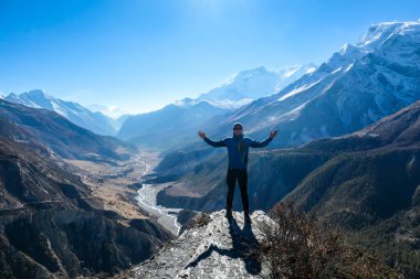 A man wearing a beanie and blue jumper, spreading his arms wide, breathing deeply the fresh mountain air. Freedom and happiness. Below Manang valley stretches in Himalayas, along Annapurna Circuit.
