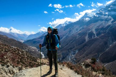 A man enjoying her trek to Tilicho Lake, along Annapurna Circuit in Nepal. In the back there are high, snow capped Himalayan peaks. Slopes are overgrown with small bushes. Exploration and discovering