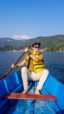 A man in yellow safety jacket, sitting in a blue boat and paddling across Phewa Lake in Pokhara, Nepal. Behind him there are high, snow capped Himalayas with Mt Fishtail (Machhapuchhare) between them